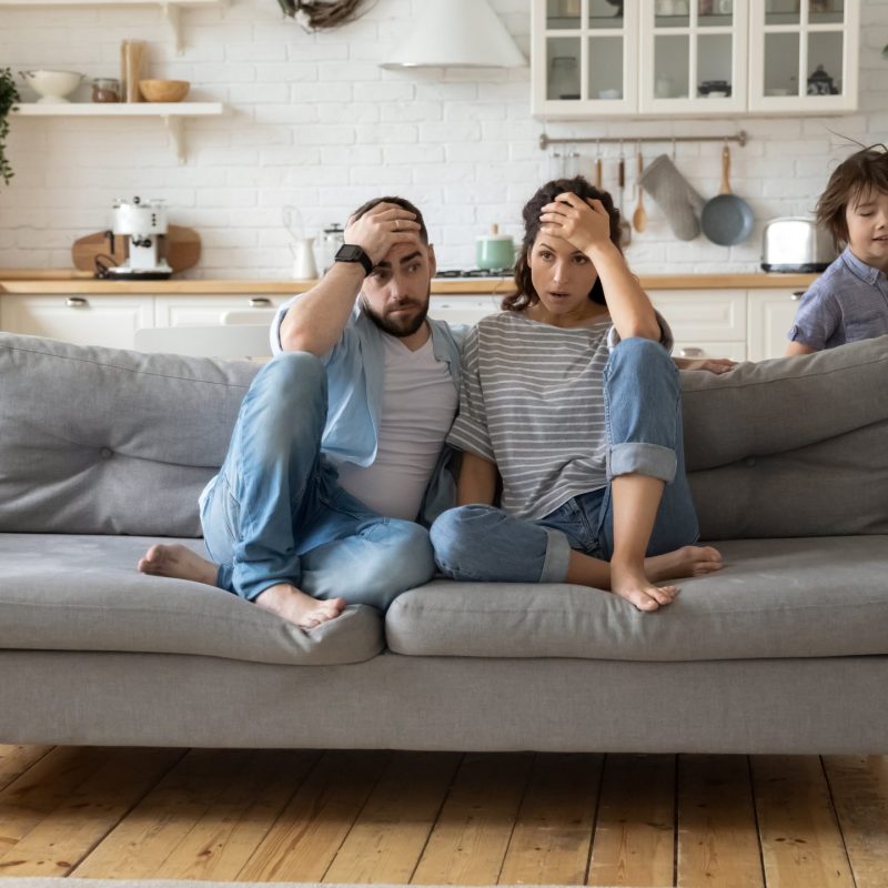 Tired mother and father sitting on couch feels annoyed exhausted while noisy little daughter and son shouting run around sofa where parents resting. Too active hyperactive kids, need repose concept