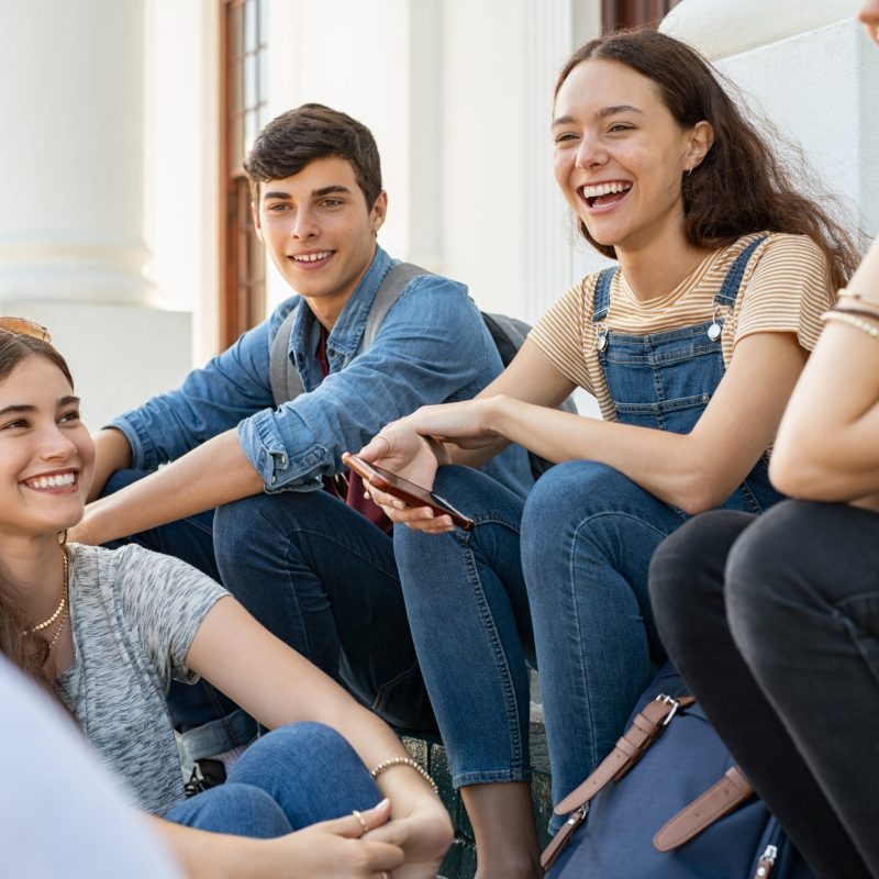 Group of happy young friends sitting in college campus and talking. Cheerful group of  smiling girls and guys feeling relaxed after university exam. Excited millenials laughing and having fun outdoor.